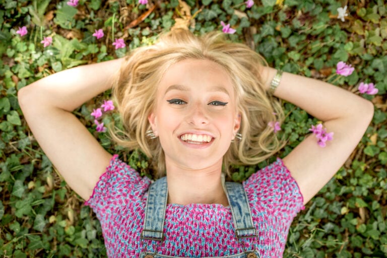 Bright blonde woman lying on grass with pink flowers, smiling.
